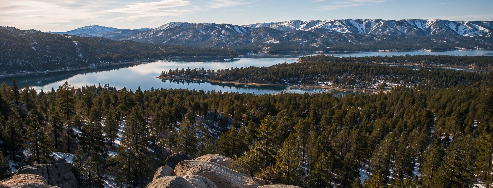 Big Bear Lake seen from a granite ridgeline, the snow-capped San Bernardino range rising behind 2,971 acres of open water at 6,742 feet.
