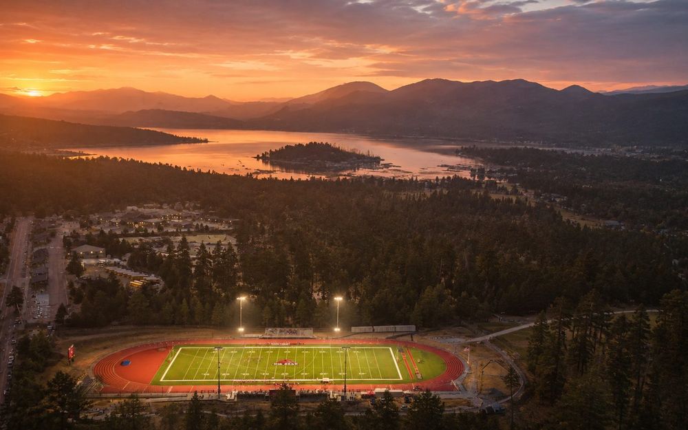 Aerial view of Big Bear Lake at sunset with a floodlit running track and athletic field in the foreground.